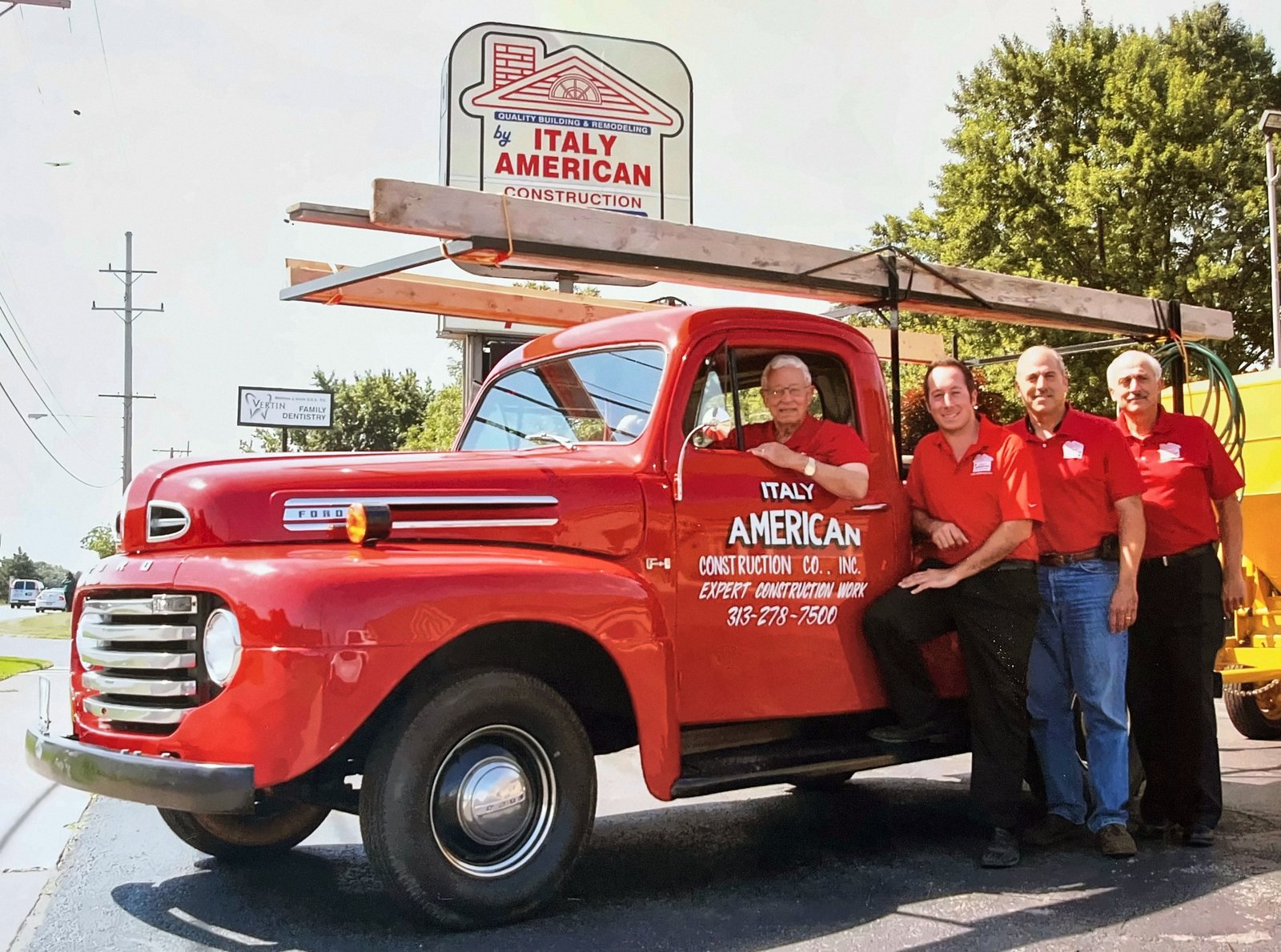 Armando Mastroianni seated in a red Ford pickup lettered Italy American Construction, with his grandson Michael and his sons Antonio and Frank standing beside it, beneath the company sign